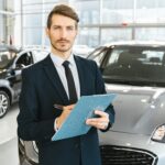 A confident car salesman in a showroom, holding a clipboard and pen.